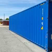 A blue 40ft shipping container sits on a paved area, surrounded by industrial buildings and equipment under a clear sky.