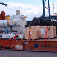 A shipping container holds a large machine and wooden crates, secured with straps at a port setting.