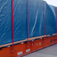 A large container covered with a blue tarp and secured with red straps, positioned at a transport facility.