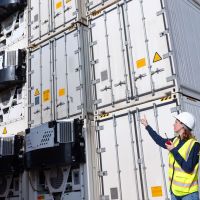 Two women in hard hats inspect refrigerated shipping containers, discussing logistics under a clear blue sky.